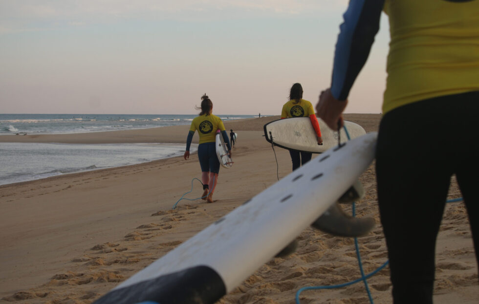 Trois surfeurs transportant des planches de surf sur une plage de sable au coucher du soleil.