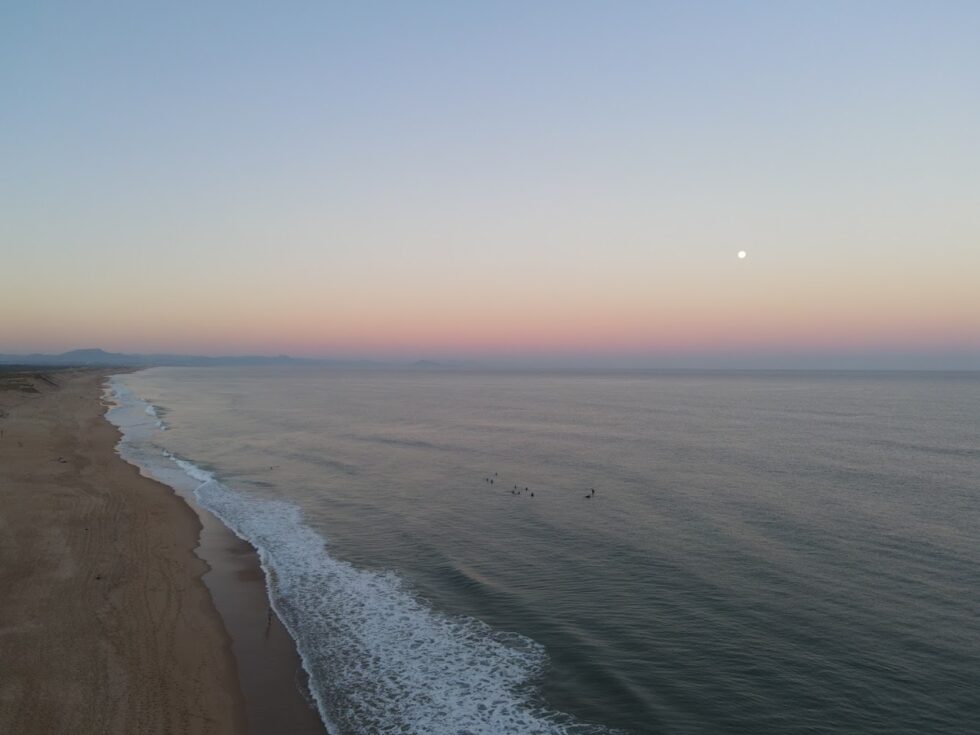 Vue aérienne de surfeurs dans l'océan au coucher du soleil, avec une plage de sable et des montagnes au loin.