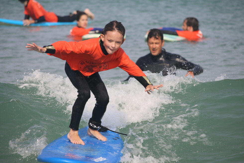 Fille en t-shirt orange Ki Surf School surfant sur une petite vague