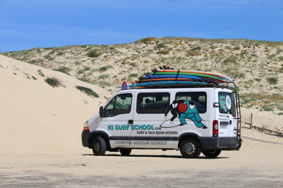 Fourgonnette blanche avec planches de surf sur le toit, garée sur une plage de sable. Logo de l'école de surf KI visible.