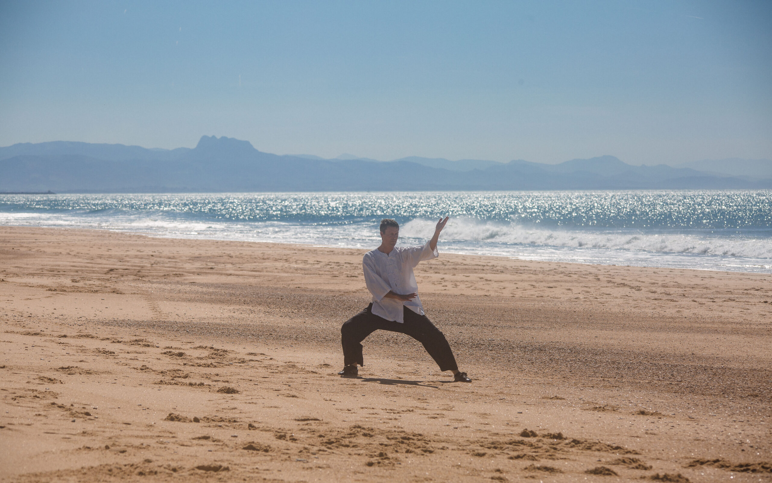 Un homme pratiquant le Tai Chi Chuan sur une plage de sable avec l'océan et les montagnes en arrière-plan.