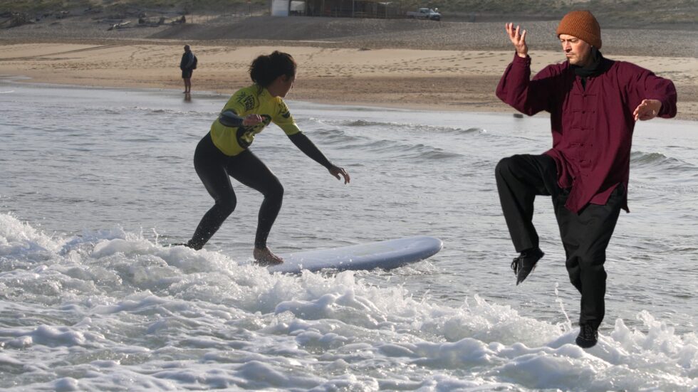 Une femme surfe tandis qu'un homme pratique le Tai Chi au bord de l'océan.