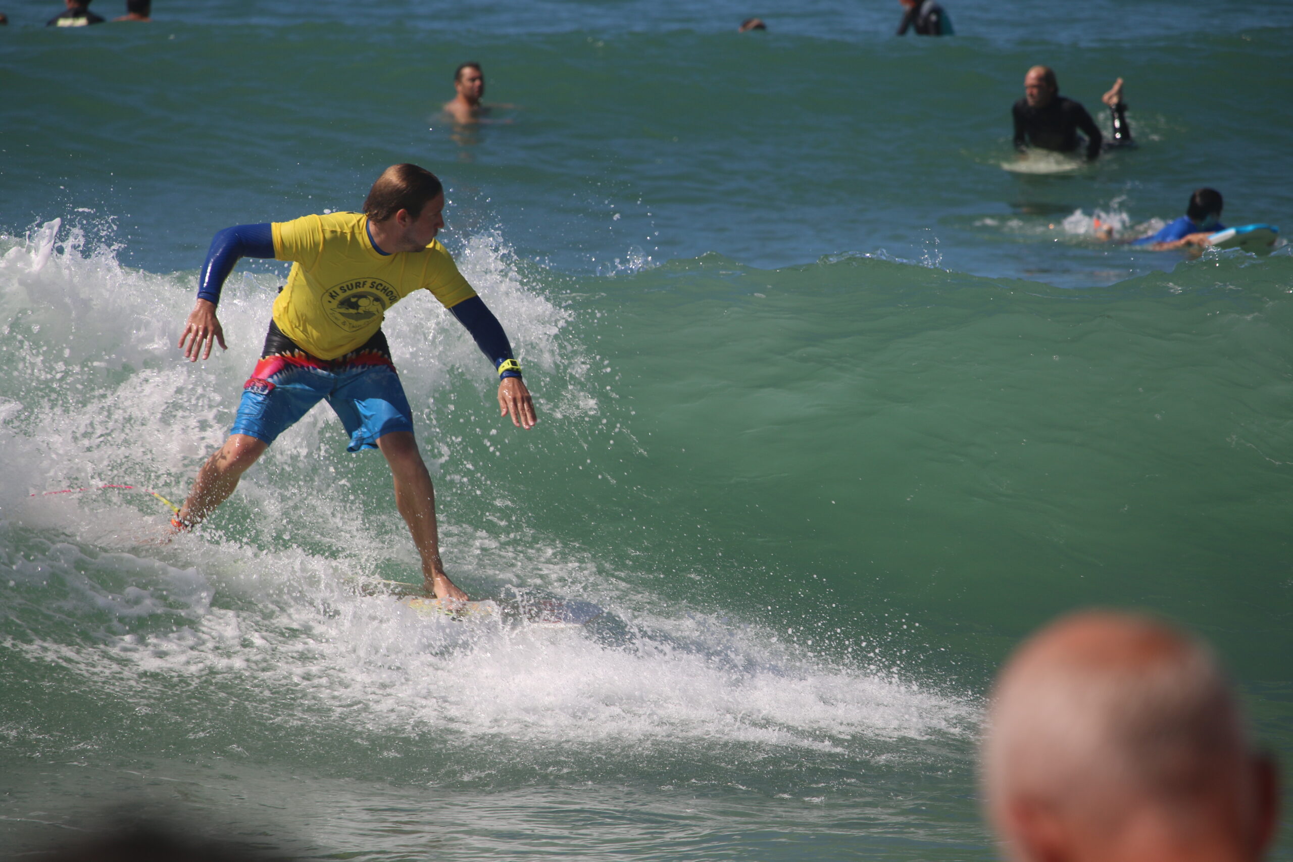 Surfeur en action sur une vague verte, école de surf visible sur le t-shirt