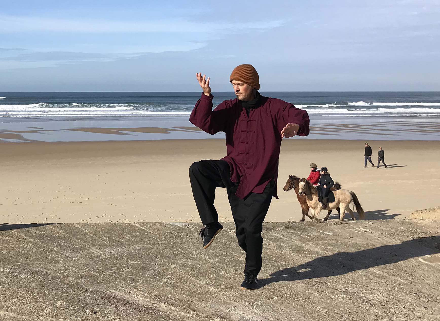 Homme pratiquant le Tai Chi sur la plage, avec des cavaliers en arrière-plan.