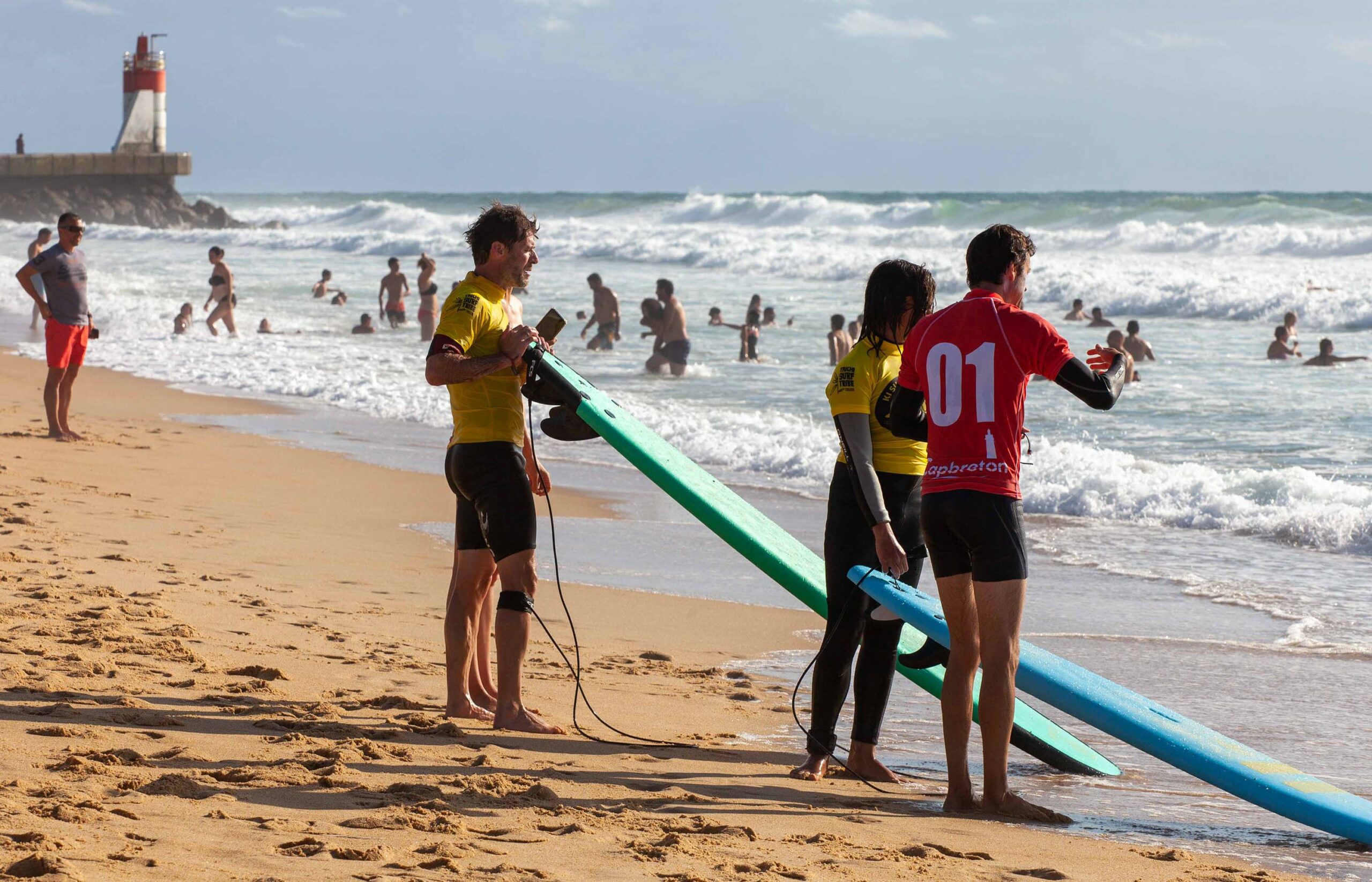 Des surfeurs se préparent sur une plage de sable avec des planches de surf et un phare au loin.